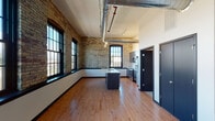 This single-floor loft space showcases a compelling blend of industrial character and modern finishes, consistent with a thoughtfully converted historic building. Original yellow brick walls run throughout, paired with large, multi-pane black-framed windows that flood the interior with natural light and offer open views of the surrounding landscape. Exposed silver ductwork traces the high ceilings, reinforcing the industrial aesthetic while adding architectural interest.

The open-concept kitchen serves as a central focal point, featuring a generous island with a light quartz countertop and undermount sink, dark espresso cabinetry extending to the ceiling, and a full suite of stainless steel appliances including a refrigerator, range, and built-in microwave. Warm hardwood flooring spans the kitchen and main living areas, contributing to an inviting atmosphere.

The bedrooms are generously proportioned, with carpet flooring, large brick-framed windows, and exposed ductwork maintaining the loft's industrial character. Two bathrooms serve the space: one featuring a soaking tub and dark vanity cabinetry, and a second equipped with a walk-in shower and accessible grab bar. Dark-framed interior doors provide cohesive, modern detailing throughout the unit.