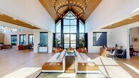 This expansive single-floor amenity center offers a thoughtfully designed collection of spaces suited for community gathering, work, and wellness. The grand lobby and main living area immediately impress with soaring vaulted ceilings clad in warm wood paneling, dramatic floor-to-ceiling windows, and an abundance of natural light. A statement chandelier and clean-lined furnishings complete the welcoming atmosphere.

Adjacent living spaces include a lounge anchored by a full-height marble-surround fireplace, exposed wood beams, and clerestory windows that fill the room with light. The dining and social area features a striking live-edge communal table, open shelving adorned with greenery, and a well-appointed kitchenette with subway tile backsplash and bar seating, with direct access to an outdoor patio.

The dedicated exercise room is outfitted with wall-to-wall mirrors, ballet barres, hardwood flooring, and direct pool-area access — ideal for fitness classes or personal workouts. A private office with corner windows and a solid wood conference table provides a quiet workspace. A small lounge nook with built-in toy storage caters to younger visitors, while a secure package and refrigerated delivery room adds everyday convenience. Light hardwood flooring flows consistently throughout, unifying the entire space.