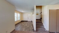 This two-story residence offers a practical and well-organized layout across both levels. The main floor welcomes you through a classic six-panel front door with glass inserts, opening into a bright living area finished with durable wood-look vinyl plank flooring. Multiple windows dressed with horizontal blinds allow natural light to fill the space, while baseboard heating units run along the walls for efficient warmth. A carpeted staircase with a wood railing leads to the upper level, conveniently positioned near the entry. Adjacent to the main living area, the kitchen is equipped with warm oak cabinetry, dark laminate countertops, and white appliances including a coil-top range, dishwasher, and refrigerator. A window above the counter adds natural light to the cooking space. The upper level features carpeted bedrooms with baseboard heating and windows overlooking the surroundings. Two bathrooms serve the home — one featuring a full soaking tub and shower combination, and another with a standalone walk-in shower stall, a wood-framed medicine cabinet mirror, and a single-sink vanity with under-cabinet storage. A dedicated utility room houses the boiler and laundry hookups. Textured ceilings are found throughout, and consistent neutral wall tones give the home a clean, cohesive appearance.