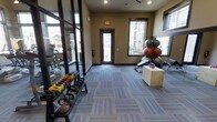 This fitness center is a thoughtfully designed, single-floor workout facility that provides a comprehensive exercise environment for its users. The space features a neutral color palette with warm beige walls, dark-trimmed windows, and patterned blue-gray carpet tiles that lend the room a clean, modern aesthetic.

Natural light flows generously through multiple large windows and a glass-paneled door, offering views of the surrounding exterior and creating an open, inviting atmosphere. A full-length mirrored wall along one side visually expands the space while allowing users to monitor their form during workouts.

The facility is well-stocked with a diverse range of equipment. A tiered dumbbell and kettlebell rack lines one wall, offering a wide selection of free weights for strength training. On the opposite side, a rack of stability and medicine balls sits alongside a workout bench and a plyo box, catering to functional fitness and conditioning exercises. Additional cardio and strength machines are visible beyond the mirror, indicating a broader equipment offering throughout the space.

Recessed ceiling lighting supplements the natural light, ensuring the space remains well-lit at all hours. An emergency exit door and clearly marked safety features reflect a practical, safety-conscious design. Overall, this fitness center delivers a functional and well-organized environment suited for a variety of training styles.