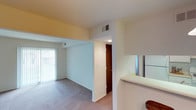 This single-floor residential space is designed with an open, flowing layout that connects the main living area to the kitchen, creating a cohesive and functional floor plan. The living area is finished with neutral-toned carpeting and bright white walls, lending a clean and airy atmosphere throughout. A large sliding glass door fitted with vertical blinds allows generous natural light to fill the space while providing access to the outdoors.

The kitchen is partially open to the living area, separated by a half-wall counter that serves as a natural divider while maintaining the open feel of the space. A full-size refrigerator is visible within the kitchen, which features light-colored cabinetry and tile flooring, offering a practical and efficient workspace.

A centrally located interior door, framed in warm wood tones, leads to an adjacent room or hallway, suggesting a well-organized layout with clear separation between living and private areas. HVAC vents are integrated into the ceiling, ensuring year-round climate comfort. The overall design emphasizes simplicity and functionality, with neutral finishes and consistent lighting throughout that make the space feel welcoming and move-in ready.