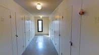 This single-floor hallway is a functional, well-organized corridor lined with multiple white doors along both sides, suggesting access to several individual rooms or storage units. The doors are uniform in design, finished in white with gold-toned hardware, lending a clean and consistent aesthetic throughout the space. The corridor is illuminated by globe-style ceiling fixtures that cast even, warm light across the length of the hall, complementing the neutral cream-toned walls and textured ceiling. The flooring consists of smooth vinyl tiles in a muted grey-blue tone, contributing to the utilitarian character of the space. At the far end of the hallway, a narrow vertical window with a dark wood frame allows a measure of natural light to filter in, brightening the terminal end of the corridor. A baseboard heater is visible beneath the window, indicating in-unit climate control. The overall ambiance is orderly and practical, with a straightforward layout designed to maximize accessibility. The space reflects a no-frills, efficient design well-suited for multi-room residential or institutional use.