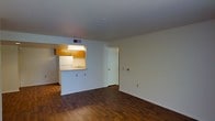 This single-floor unit features an open-concept layout that connects the living area to the kitchen through a pass-through window, creating a sense of connectivity while maintaining defined spaces. The living area is finished with rich, dark hardwood flooring that extends throughout, adding warmth and visual depth to the space. Crisp white walls provide a neutral backdrop and contribute to the bright, airy atmosphere of the room.

The kitchen is accessible via a pass-through opening and is outfitted with warm honey-toned wood cabinetry, an under-cabinet light fixture, a white refrigerator, and a sink positioned beneath the pass-through counter. The kitchen's compact design makes efficient use of the available space while remaining clearly visible and connected to the main living area.

A white interior door off the main living space leads to an additional room or hallway, suggesting further functional areas beyond what is immediately visible. Recessed ventilation in the ceiling and standard electrical outlets along the walls reflect practical, straightforward construction. Overall, the unit offers a clean, functional environment suited for comfortable everyday living.