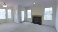This two-story home offers a welcoming living space highlighted by a well-appointed fireplace framed in warm wood trim with a dark surround, serving as the room's focal point. The living area features neutral-toned carpeting and light-colored walls that create a clean, airy atmosphere. Multiple windows, including a glass-paneled exterior door, flood the space with natural light from several angles, enhancing the open feel of the room. Recessed ceiling lighting provides additional illumination, while the layout offers generous floor space suitable for a variety of furniture arrangements. The exterior door provides convenient access to the outdoors, adding to the functionality of the main living area. The overall design reflects a traditional residential style with practical finishes throughout, making it a comfortable and versatile living environment across both floors.