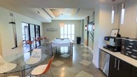This single-floor recreational lounge is designed with a contemporary aesthetic, combining functionality and style in a well-appointed layout. The space is anchored by a sleek white pool table with polished chrome legs, positioned beneath a decorative pendant light that adds a warm, focused glow to the area. The ceiling features a tray design with recessed lighting, contributing to the room's refined character and sense of vertical dimension.

The flooring is a striking combination of hardwood and inlaid tile in a geometric pattern, adding visual texture underfoot. Natural light enters through shuttered windows along one wall, brightening the neutral gray-toned walls and creating an airy atmosphere. A casual seating area with a round glass table and modern orange accent chairs provides a social gathering spot, while bar-height seating with orange stools lines one side of the room.

A large flat-screen TV is mounted on a decorative accent wall, complemented by stylized vertical wall art and greenery. A small refreshment station with a beverage appliance is conveniently positioned nearby, enhancing the lounge's entertainment-ready functionality. The overall ambiance is modern, clean, and inviting.