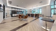 This single-floor amenity space is thoughtfully designed to offer a variety of social and recreational environments within one cohesive layout. The main lounge is expansive and open, featuring light wood-toned flooring accented by dark marble inlays, recessed and pendant lighting, and a striking geometric black feature wall. Seating options range from leather and chrome dining sets to bar-height chairs positioned along floor-to-ceiling windows that frame lush tropical courtyard and pool views, filling the space with abundant natural light.

A dedicated media lounge area includes a large multi-screen wall display, complemented by swivel accent chairs and a small-group seating arrangement. An adjacent banquette zone offers continuous teal upholstered bench seating with rounded ottomans, ideal for casual gatherings. Near the entrance, a sculptural art piece on a pedestal adds a refined focal point, while sheer curtains soften the natural light along the window walls.

The floorplan also incorporates a fitness area equipped with exercise machines and a game room with a pool table. Throughout, the design maintains a clean, contemporary aesthetic with consistent flooring, neutral walls, and curated furnishings that create a welcoming and functional communal atmosphere.