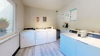 This shared laundry facility at 4729 Manzanita Ave in Carmichael, California, offers a clean and efficiently organized space designed for everyday convenience. The room features light wood-style flooring and neutral-toned walls that create a bright, welcoming atmosphere. Multiple commercial-grade front-loading washers and top-loading dryers are arranged along the walls, providing ample capacity for residents. A wall-mounted payment kiosk offers a modern, cashless method for operating the machines. Posted laundry room rules, instructional guides, and a cork bulletin board keep users informed and help maintain an orderly environment. A large window along one wall allows natural light to fill the space while offering a view of the surrounding greenery outdoors. The entry door is solid and clearly marked with occupancy guidelines. The room maintains a tidy, well-maintained appearance throughout, with a dark baseboard accent providing a clean visual boundary between the flooring and walls. Overall, this laundry room is a practical and accessible shared amenity, thoughtfully equipped to serve the needs of the surrounding residential community.