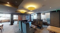This thoughtfully designed amenity lounge occupies a single floor and offers a variety of distinct zones to accommodate different activities and social settings. The space is anchored by a comfortable lounge area furnished with plush, high-back wingback chairs in rich velvet upholstery, arranged around a live-edge wood coffee table. Signature pendant and oversized drum-shade ceiling fixtures cast a warm, ambient glow throughout.

A semi-private nook with built-in banquette seating and wooden dining tables provides a quieter retreat, while a dedicated dining area offers additional seating with woven-seat chairs and floating shelves lined with small decorative plants. A well-appointed kitchenette features warm wood cabinetry, a dark stone countertop, and a herringbone tile backsplash, adding both function and style.

The space also includes a foosball table for recreation, curated wall art, and nature-inspired photography displayed throughout. Textured wall panels in light wood tones and bold chevron-patterned wallcoverings create visual interest and define each zone. Large floor-to-ceiling windows flood the interior with natural light and frame expansive city views, while a glass door leads directly to an outdoor terrace. The patterned carpet flooring ties the entire space together with a cohesive, contemporary aesthetic.