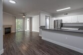 This single-floor residence is designed with a clean, contemporary aesthetic throughout. The open-concept kitchen serves as a central feature, showcasing white shaker-style cabinetry, dark gray countertops, and a full suite of stainless steel appliances, including a French door refrigerator, microwave, and gas range. A peninsula with matching countertop provides additional workspace and separation from the adjacent living areas. Wood-look vinyl plank flooring runs throughout the main living spaces, adding warmth and visual continuity.

The living room is a comfortable and well-lit space, highlighted by crown molding, recessed lighting, a ceiling fan, and a tiled fireplace as a focal point. Sliding glass doors open to an outdoor area, allowing natural light to flow in and offering views of surrounding greenery.

The bedrooms are carpeted and feature neutral-toned walls, ceiling fans, and windows with horizontal blinds that admit natural light while maintaining privacy. Closet access is present in at least one bedroom.

The bathroom is finished in a similarly modern style, with a white vanity, gray countertop, a backlit mirror, and a tub-shower combination with tiled surround. Wood-look flooring extends into this space, maintaining a cohesive feel throughout the unit.