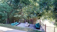 The Firepit Lounge is a well-appointed outdoor deck space designed for relaxed gathering and social enjoyment. The deck is constructed with grey composite wood planking and is enclosed by a sturdy dark metal railing system supported by wood posts, providing both safety and an open feel that preserves the surrounding natural views.

At the heart of the space sits a rectangular, modern gas fire pit with a glass wind guard and lava rock fill, serving as the natural focal point for conversation and evening gatherings. Surrounding the fire pit, multiple modular wicker seating units with vibrant teal cushions are arranged to encourage group interaction, comfortably accommodating several guests.

The lounge is embraced by a mature tree canopy that provides generous natural shade, creating a cool and serene atmosphere. A natural stone retaining wall borders one side of the deck, adding a rustic architectural element that complements the adjacent building's stone facade. Potted planters add touches of greenery at the deck perimeter.

Stair access connects the deck to surrounding pathways, and the open railing design allows unobstructed sightlines into the lush wooded landscape beyond. The combination of natural materials, abundant greenery, and thoughtful seating arrangements makes the Firepit Lounge an inviting outdoor venue suited for both casual relaxation and social events.