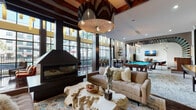 This sophisticated amenity lounge is a masterclass in contemporary design, blending dramatic architectural elements with carefully curated interiors. The space is anchored by soaring vaulted ceilings adorned with rich, dark wood beam detailing and an oversized sculptural pendant light that serves as a striking focal point. Floor-to-ceiling black-framed windows line the walls, flooding the interior with abundant natural light and offering views of the surrounding outdoor spaces.

The main lounge area features a sleek, modern fireplace, plush neutral-toned seating, and an organic live-edge coffee table that adds a natural, artistic touch. A colorful area rug grounds the seating arrangement, adding warmth and vibrancy to the space.

Adjacent to the lounge, a dedicated billiards area showcases a handsome wood-framed pool table with a turquoise felt surface, lit by a contemporary geometric chandelier. A built-in bench seating nook and decorative tile accent wall add character to this social zone.

A wet bar and café counter with bar seating is elegantly positioned beneath a curved architectural detail, complemented by open shelving for décor and essentials. Rich, dark hardwood floors flow seamlessly throughout the space.

The entry corridor features padded leather wall panels, curated artwork, and stylish drum stools, creating a welcoming and polished arrival experience.