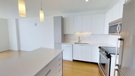 This single-floor, one-bedroom, one-bathroom unit offers a well-designed layout that maximizes its footprint with thoughtful, modern finishes. The kitchen serves as a standout feature, showcasing flat-front white cabinetry that extends to the ceiling for ample storage. A subway tile backsplash adds subtle texture against the otherwise clean, minimalist aesthetic. Stainless steel appliances, including a gas range and dishwasher, provide both style and functionality. A central island with a white quartz countertop offers additional prep space and casual seating potential. A sleek pendant light hangs above, providing focused task lighting that complements the recessed ceiling fixtures. Light hardwood-style flooring runs throughout, adding warmth to the bright, white-dominated palette. The overall ambiance is crisp, clean, and contemporary, with a neutral color scheme that creates a versatile backdrop. The open kitchen layout connects naturally to the living areas, promoting an airy, cohesive flow throughout the space.