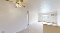 Located at 1659 Munroe Falls Ave in Cuyahoga Falls, Ohio, this single-floor unit offers three bedrooms, one bathroom, and a kitchen within a compact, efficiently arranged floor plan. The living area features neutral-toned carpet, a textured ceiling, and a ceiling fan with light fixture, creating a simple and functional gathering space. A large window allows natural light to fill the room, offering views of the surrounding neighborhood. The layout flows openly between the living area and kitchen, which is equipped with wood-toned cabinetry, a coil-burner electric range, and a full-size refrigerator, all set on easy-care hard flooring. The generously sized primary bedroom is the standout room, featuring plush carpet, a wall-mounted air conditioning unit, baseboard heating, and abundant natural light from a large sliding window. Additional bedrooms are carpeted and each include louvered bi-fold closet doors, providing practical storage. The shared full bathroom features light wood-look flooring, a tub-shower combination with white surround, a toilet, and a vanity with countertop. Baseboard heating is present throughout, and the overall palette of white walls and light finishes gives the space a clean, bright character.