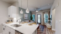 Located at 211 Satori Way on scenic Johns Island, South Carolina, this thoughtfully designed single-floor residence combines modern finishes with warm, inviting aesthetics. The open-concept layout seamlessly connects the kitchen and living area, creating an ideal space for both everyday living and entertaining. The kitchen features quartz countertops, a stainless steel refrigerator, white cabinetry with black hardware, and a central island illuminated by stylish pendant lights. A built-in wine rack adds a refined touch. The living area is anchored by a sage green accent wall, light hardwood-style flooring, and sliding glass doors that invite abundant natural light and outdoor access. A ceiling fan adds comfort overhead. A secondary sitting or media area offers a clean, minimalist layout with wall-mounted TV and ample natural light through plantation-shuttered windows. The bedroom is a serene retreat highlighted by a dramatic botanical-print accent wall, large windows with garden views, and a ceiling fan. The bathroom features a tiled tub-shower combination, white vanity with modern fixtures, and hardwood-style flooring. Completing the home is a dedicated in-unit laundry area with full-size washer and dryer, and convenient overhead shelving storage.