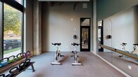This well-appointed fitness facility offers a thoughtfully designed single-floor layout that caters to a wide range of workout styles. The space is divided into distinct functional zones, beginning with a dedicated spin studio housing stationary cycling bikes, a wall-mounted mirror with a ballet barre, and a flat-screen TV — ideal for guided cycling sessions or low-impact training. Exposed concrete columns and dark-painted beams lend the studio an industrial aesthetic, complemented by a muted, teal-toned color palette on the accent walls. An interior glass door connects the spin studio to the main workout area, ensuring seamless flow between spaces. The primary fitness floor is equipped with an organized kettlebell rack spanning a range of weights, stability balls, dumbbells, and additional cardio equipment. A large floor-to-ceiling window on the main floor floods the space with natural light and offers open views of the surrounding urban streetscape. A full-length wall mirror amplifies the sense of space and provides visual feedback during workouts. The carpeted flooring throughout the facility offers a clean, comfortable surface suitable for stretching and floor exercises. Together, the industrial design elements, generous natural light, and diverse equipment selection make this a versatile and inviting fitness environment.