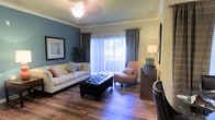 This single-story living space combines classic architectural details with a comfortable, well-appointed interior. The living room serves as the focal point of the home, showcasing rich hardwood flooring that runs throughout and adds warmth to the overall aesthetic. Crown molding frames the ceiling, lending an elegant, polished finish to the room.

A striking teal accent wall provides a bold contrast to the surrounding neutral tones, creating visual interest and depth. A ceiling fan with a light fixture anchors the ceiling, contributing both function and style. Natural light pours into the space through a large sliding glass door dressed with sheer white curtains and layered dark drapery panels, offering both brightness and privacy options.

The living area is thoughtfully arranged with a plush sofa adorned with decorative pillows, a tufted leather storage ottoman as a centerpiece, and an accent chair that complements the overall color palette. A wooden console hosts a table lamp, adding ambient lighting. Framed wall art and indoor greenery contribute finishing touches that enhance the inviting, curated ambiance of this well-designed space.