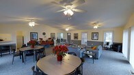 This single-floor space is designed to accommodate both casual dining and relaxed lounging within one open, flowing layout. The room is divided into two functional zones that coexist comfortably, creating a versatile gathering environment. The dining area, situated toward the front, features round tables paired with black chairs, offering ample seating for groups. Decorative red poinsettias serve as centerpieces, adding a warm, welcoming touch. The lounge area toward the rear is furnished with gray upholstered sofas and modern glass-topped coffee tables, creating a comfortable setting for socializing. The space is finished with neutral yellow and gray two-tone walls accented by framed artwork, contributing to a clean, inviting ambiance. Multiple ceiling fans with integrated lighting fixtures are evenly distributed across the ceiling, ensuring consistent airflow and illumination throughout. Vertical blinds on the windows allow for adjustable natural light. A secondary room visible in the background suggests additional utility or office space. The carpeted flooring ties the entire area together, enhancing the comfort and cohesion of the overall space.