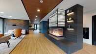 This contemporary amenity lounge is thoughtfully designed to offer a dynamic and inviting community experience across a single, well-appointed floor. At the heart of the space stands a dramatic three-sided gas fireplace crowned by a sculptural stacked-panel hood, flanked by dark textured accent walls with integrated shelving and ambient lighting, creating a striking focal point upon entry. Warm wood-look flooring and a rich wood-paneled ceiling add natural warmth throughout.

The lounge flows seamlessly into multiple distinct zones. A sitting area features designer lounge chairs and a sofa set against a dimensional brick accent wall, bathed in natural light from expansive floor-to-ceiling glass walls that frame outdoor neighborhood views. A dedicated dining nook with mid-century modern chairs and a round table sits beneath a sculptural pendant light, with curated wall art adding an artistic touch.

The wet bar area is a showpiece of its own, boasting a dramatic veined stone countertop island with bar seating, globe pendant lighting, and a vibrant neon heartbeat art installation against an exposed brick backdrop. A game room with a full-size pool table, access to an outdoor terrace, and additional lounge seating rounds out the entertainment offerings. The space is completed by a curved sectional lounge area wrapped in floor-to-ceiling glazing, blurring the line between indoors and the outdoor patio beyond.