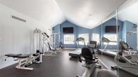 This dedicated fitness space is thoughtfully designed to support a comprehensive workout experience within a single-floor layout. The room features a striking vaulted ceiling that rises to a sharp peak, lending a sense of openness and airiness to the space. The walls are painted in a cool slate blue on the accent sides, contrasting with crisp white on the upper portions and ceiling, creating a clean, energizing aesthetic. Recessed lighting and a textured ceiling complement the overall design.

The rubber-floored space is well-equipped for both cardiovascular and strength training. Cardio equipment includes treadmills and a recumbent stationary bike, while the strength training area features a multi-function cable machine, a weight bench, and a dumbbell rack with a full set of free weights. Stability balls and resistance bands add versatility to available workouts. A large flat-screen television is mounted on the wall, and a full-length mirror panel along one wall visually expands the space while supporting form monitoring during exercise. Multiple windows fitted with blinds allow natural light to filter in, and a water cooler provides convenient hydration. The space also includes a dedicated dumbbell storage area and wall-mounted fitness accessories.