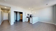 This single-floor space showcases a clean, modern design highlighted by an open-concept living and kitchen area. The kitchen is outfitted with white cabinetry, a white subway tile backsplash, and a full suite of stainless steel appliances, including a refrigerator, electric range, and microwave. A peninsula with a built-in sink extends into the living area, offering additional counter space and serving as a natural divider between the kitchen and the main living space. Three sleek pendant lights hang above the peninsula, adding a contemporary touch to the space. The living area features smooth gray luxury vinyl plank flooring that runs throughout, complementing the neutral light gray walls and white trim. Recessed lighting in the kitchen ceiling supplements the pendant fixtures, ensuring the space is well-lit. A doorway visible from the main living area leads to an additional room with darker flooring, suggesting a distinct transition in zones. The high ceilings contribute to an airy, open feel, making the overall space feel spacious and inviting despite its single-story footprint.