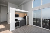 This thoughtfully designed single-floor unit blends contemporary style with functional living. The open-concept kitchen serves as a central feature, outfitted with warm wood-toned upper cabinetry, sleek grey lower cabinets, a subway tile backsplash, and a full suite of stainless steel appliances including a range, refrigerator, and over-the-range microwave. Light countertops provide ample workspace, while a sliding glass door opens directly onto a private balcony enclosed with a decorative perforated screen panel, inviting natural light and outdoor access. The soaring ceiling height above the kitchen adds a striking sense of vertical space. The living area benefits from large, floor-to-ceiling windows that flood the room with abundant natural light, casting dramatic geometric shadows across the light wood-tone flooring that runs throughout the unit. A sliding barn-style door adds a modern industrial accent in the entryway corridor. The bathroom is cleanly appointed with a combination tub and shower, a vanity with a white countertop, modern grey cabinetry with gold hardware, and a large mirror above. The overall design palette is neutral and contemporary, emphasizing clean lines, natural light, and cohesive finishes throughout.