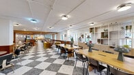 This expansive single-floor amenity space blends retro-inspired aesthetics with modern functionality across a thoughtfully designed open floor plan. The standout architectural feature is the bold black-and-white terrazzo checkerboard flooring that anchors the main open area, complemented by warm wood wainscoting running along the walls and creating a cohesive mid-century modern atmosphere throughout.

The space is organized into several distinct yet interconnected zones. The main lounge area offers a variety of seating arrangements, including banquette-style booths with colorful upholstery, lounge chairs, and café-style tables, making it ideal for both casual gatherings and individual work. A dedicated relaxation nook features a richly upholstered channeled leather sofa beneath a warm slatted wood ceiling, with a large-format game board adding a playful touch and natural light filtering in through multiple windows.

A private conference room provides a more formal setting, appointed with a substantial conference table, plush velvet seating, a wall-mounted display screen, and eye-catching patterned wallcovering. An open shelving unit serves as a stylish room divider, while integrated lighting throughout ensures a well-lit, welcoming environment. The overall ambiance is warm, eclectic, and highly functional—designed to foster community and productivity alike.