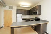 This single-floor unit offers a well-organized layout with distinct living areas designed for comfortable everyday living. The kitchen is a standout feature, outfitted with dark espresso-toned cabinetry, stainless steel appliances including a refrigerator, electric range, and microwave, and an extended countertop that doubles as a breakfast bar. Light wood-look flooring runs throughout the kitchen and main living areas, lending the space a clean, contemporary feel. The open configuration of the kitchen allows for easy flow into the adjoining living area, making the space feel open and connected. The bedrooms are carpeted for added comfort and are finished with neutral walls and bold black baseboards that provide a modern contrast. One bedroom benefits from a large window offering elevated views of the surrounding cityscape, filling the room with natural light. The bathroom is clean and functional, featuring a full-size soaking tub with an overhead shower, white surround walls, and the same light wood-look flooring found throughout the unit. The overall design is straightforward and modern, with a neutral color palette that provides a versatile backdrop suitable for a variety of interior styles.