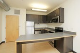 This single-floor unit offers a well-organized layout with distinct living areas designed for comfortable everyday living. The kitchen is a standout feature, outfitted with dark espresso-toned cabinetry, stainless steel appliances including a refrigerator, electric range, and microwave, and an extended countertop that doubles as a breakfast bar. Light wood-look flooring runs throughout the kitchen and main living areas, lending the space a clean, contemporary feel. The open configuration of the kitchen allows for easy flow into the adjoining living area, making the space feel open and connected. The bedrooms are carpeted for added comfort and are finished with neutral walls and bold black baseboards that provide a modern contrast. One bedroom benefits from a large window offering elevated views of the surrounding cityscape, filling the room with natural light. The bathroom is clean and functional, featuring a full-size soaking tub with an overhead shower, white surround walls, and the same light wood-look flooring found throughout the unit. The overall design is straightforward and modern, with a neutral color palette that provides a versatile backdrop suitable for a variety of interior styles.