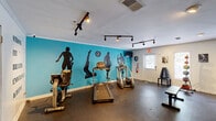 Located at 301 W Vandalia Rd in Greensboro, NC, this dedicated exercise room delivers a focused and energizing fitness environment within a single, efficiently utilized space. The room is immediately distinguished by a bold teal accent wall adorned with large black silhouettes of athletes in dynamic poses, creating a motivating visual atmosphere reinforced by bold typographic wall decals reading "Strong, Fit, Breathe, Endurance, Achieve." Track lighting mounted on the white ceiling provides bright, directed illumination throughout the space. The flooring consists of durable rubber fitness matting, well-suited for heavy equipment and high-impact activity. Cardio equipment includes a treadmill, an elliptical trainer, and a stationary bike, while a rack of dumbbells, a set of medicine balls, and a flat workout bench cater to strength training. A large floor-to-ceiling mirror along one wall visually expands the room and supports form-checking during exercise. Natural light enters through multiple windows flanking the entry door, contributing to an open and welcoming feel. A wall-mounted television provides entertainment during workouts, and a fire extinguisher and exit signage ensure safety compliance. The space is thoughtfully designed to offer a comprehensive workout experience in a compact, purpose-built setting.