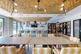 Located at 960 Greene St in Ann Arbor, Michigan, this contemporary amenity space spans a generous single floor, offering a well-appointed communal living area designed for both relaxation and socializing. The interior is immediately distinguished by a striking textured wood-panel ceiling that anchors the space with warmth and visual depth, complemented by bold black pendant lighting throughout.

The living room area features multiple seating zones, including plush sofas and accent chairs arranged around coffee tables, creating an inviting lounge atmosphere. A communal dining area with a long wooden table and patterned upholstered chairs sits adjacent to a bar-height counter with additional seating. A curated shelving display and a flat-screen TV add to the social amenities, while a full-size pool table serves as a centerpiece for recreation.

Large floor-to-ceiling sliding glass doors flood the interior with natural light and provide seamless access to the outdoor terrace. Outside, a resort-style swimming pool is framed by rows of lounge chairs and a wood-slatted pergola with string lighting. A basketball hoop and additional seating areas with fire features enhance the outdoor experience. The building's modern facade — featuring bold yellow and gray paneling with private balconies — rises above the pool deck, offering a dynamic architectural backdrop to this vibrant amenity space.