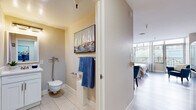 Located in Oakland, CA, this thoughtfully designed one-bedroom, one-bathroom residence offers a compact yet functional layout across a single floor. The main living area serves as the heart of the space, combining sleeping, dining, and living functions in an open, airy arrangement. Light hardwood flooring runs throughout, lending warmth and continuity to the room. Large floor-to-ceiling windows flood the space with natural light and provide views of the surrounding urban greenery and balcony railing, creating a bright and inviting atmosphere.

The sleeping area is generously proportioned, accommodating a full-sized bed alongside a dining table with blue upholstered chairs and additional seating. Coastal-themed artwork adorns the walls, contributing to the space's calm, relaxed aesthetic. A compact kitchenette is neatly integrated into the space, featuring white cabinetry with a subway tile backsplash, a microwave, and a full-sized stainless steel refrigerator—providing practical functionality without interrupting the open layout.

The bathroom is finished with large-format light tile flooring, a wall-mounted toilet, a white vanity with under-cabinet storage, and a tiled shower area with decorative mosaic accents. A nautical-inspired painting adds a cohesive decorative touch. The overall ambiance of the space is clean, modern, and serene, with a coastal color palette of whites, beiges, and blues throughout.