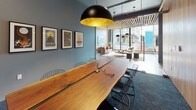This thoughtfully designed conference room combines raw natural materials with contemporary aesthetics to create a productive and visually engaging meeting space. At the heart of the room sits a commanding live-edge solid wood table, its organic form and warm grain providing a striking focal point that seats a generous number of participants. Sleek wire-frame chairs surround the table, balancing the table's rustic character with an industrial edge.

The room features two distinct wall treatments: one wall is clad in light vertical wood paneling, adding warmth and texture, while the opposing wall is finished in a cool slate-blue-grey tone and adorned with a curated row of framed concert posters, lending the space a creative, urban personality. A wall-mounted flat-screen display serves functional presentation needs on the far end.

Overhead, bold black dome pendant lights with gold-leaf interiors cast a warm, focused glow over the workspace. Integrated cable management is built into the table surface, accommodating modern connectivity needs. Dark charcoal carpet grounds the space underfoot.

Glass partitioning on one side opens the conference room to an adjacent lounge area with plush seating and floor-to-ceiling windows, allowing natural light to filter in and providing views of the surrounding exterior — connecting the intimate meeting environment to a broader, light-filled communal setting.