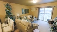 This single-floor space centers around a warm and inviting living room designed for comfort and functionality. The living room features neutral-toned carpeting that complements the soft, beige walls, creating a cohesive and calming atmosphere throughout. A large sliding glass door dominates one wall, allowing natural light to pour into the space while providing direct access to an outdoor area, offering views of the surrounding exterior.

The living area is generously sized, comfortably accommodating a large sectional-style sofa dressed with layered throw pillows and blankets in coordinating teal, cream, and earth tones. A rustic wooden coffee table serves as a functional centerpiece. Wall art above the sofa adds a nature-inspired focal point, while a floor lamp in the corner provides additional ambient lighting alongside the flush-mount ceiling fixture overhead.

The overall ambiance of the space is relaxed and welcoming, with a neutral color palette accented by subtle decorative touches. The layout is practical and open, allowing for easy movement and a clear sightline from the main seating area to the outdoor access point, making the space feel connected and airy.