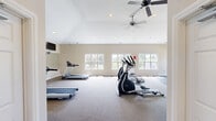 Located at 100 Crimson Rose Dr in Huntsville, Alabama, this community fitness center offers a well-equipped, single-floor workout facility designed to support a full range of exercise routines. The space features soaring vaulted ceilings that lend an open, airy feel, complemented by recessed lighting and dual ceiling fans that maintain comfortable air circulation throughout. Neutral-toned walls and carpeted flooring in the main cardio area create a clean, welcoming environment, while a dedicated rubber-floored zone provides a stable surface for strength training. Multiple windows along the walls invite abundant natural light and views of the surrounding outdoors, enhancing the overall atmosphere. The cardio section is equipped with treadmills, elliptical trainers, stair climbers, and a recumbent bike, while a multi-function cable machine and a dumbbell rack with a varied weight selection round out the strength training offerings. Wall-mounted televisions are positioned for easy viewing during workouts. A large floor-to-ceiling mirror along one wall visually expands the space and supports proper form. Additional amenities include a dual water fountain, a first aid station, and an AED unit, ensuring safety and convenience for all users.