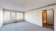 Located at 414 Erie St SE in Minneapolis, Minnesota, this two-bedroom, one-bathroom unit offers a practical and well-organized single-floor layout. The living area is generously proportioned, featuring wall-to-wall carpet, a textured ceiling, and a large window with blinds that allows natural light to fill the space. A baseboard heating unit and a wall-mounted AC unit ensure year-round comfort. The kitchen is efficiently designed with warm wood cabinetry, light countertops, tile flooring, and white appliances including a refrigerator, electric range, and dishwasher, along with a double sink beneath additional upper cabinets. Both bedrooms are carpeted and feature natural light from windows with blinds and baseboard heating. The primary bedroom offers generous proportions, while the second bedroom is equipped with a wide set of double bi-fold closet doors, providing substantial storage. The full bathroom includes a tiled tub-shower combination with a sliding glass door enclosure and a clean, functional layout. A connecting hallway with additional closet access ties the rooms together efficiently, making this a well-appointed and organized living space.