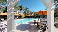 This outdoor amenity area is centered around a generously sized swimming pool with crystal-clear blue water, framed by a wide concrete pool deck designed for both relaxation and socializing. Multiple lounge chairs and chaise seating are arranged along the poolside, each shaded by vibrant orange market umbrellas that add a warm, resort-like atmosphere to the space. A pergola structure anchors a dedicated outdoor dining area, where slatted tables and cushioned chairs provide a comfortable spot to gather beneath additional umbrella shade. The surrounding landscape features mature palm trees, lush green hedgerows, and carefully maintained garden beds that create a sense of privacy and tropical ambiance. Walkways wind through the grounds, connecting the pool deck to adjacent green lawn areas and other amenities. Restroom facilities are conveniently located nearby, accessible via the property office. The overall design emphasizes open-air comfort, with thoughtful separation between active and relaxing zones. Towering trees and multi-story residential buildings visible in the background complete the setting, contributing to the community feel of this well-maintained outdoor recreation space under open, sunny skies.