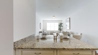 This single-floor residence offers a thoughtfully designed open-concept layout that connects the kitchen and living areas in a seamless, functional flow. The kitchen features elegant granite countertops with warm beige and gold toning, complemented by cream-colored cabinetry with classic hardware — a refined yet practical space well-suited for both everyday living and entertaining. A generous island anchors the kitchen, providing additional prep and dining space.

The living area extends naturally from the kitchen, bathed in natural light streaming through a large window that brightens the neutral, airy palette throughout. The space is finished with crisp white walls that enhance the sense of openness and calm. Tasteful décor, including a decorative mirror, framed artwork, and a lush indoor plant, adds character and warmth without overwhelming the clean aesthetic.

Seating in the living area is arranged to encourage relaxed conversation and comfort, with soft neutral furnishings that harmonize with the overall tone of the space. The cohesive design language — combining natural stone, soft textiles, and abundant light — creates an inviting ambiance that is both sophisticated and approachable throughout this well-appointed residence.