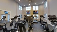 Located at 1 Mirada Dr N in Lewis Center, Ohio, this single-floor fitness center is designed to offer a well-equipped and comfortable workout environment. The space features soaring ceilings with recessed lighting and a ceiling fan, creating an open and airy atmosphere that enhances the overall exercise experience. Large, multi-pane windows line the walls at both standard and elevated heights, flooding the interior with generous natural light while providing ventilation and a sense of openness. The flooring consists of durable, textured fitness-grade material suited for high-traffic use. A variety of cardio equipment is thoughtfully arranged throughout the space, including treadmills, elliptical trainers, and stationary bikes — catering to a range of fitness preferences. Two wall-mounted flat-screen televisions are positioned prominently for easy viewing from multiple machines, allowing members to stay entertained during their workouts. The neutral color palette of warm whites and soft grays contributes to a clean, focused atmosphere. Overall, this fitness center is thoughtfully designed to balance functionality with comfort, making it an inviting space for daily exercise routines.
