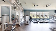 Located at 50 Greenleaf Road in Conyers, Georgia, this fitness center offers a well-appointed, single-floor exercise space designed to support a comprehensive workout experience. The room is finished with eye-catching herringbone-patterned luxury vinyl flooring in a warm wood tone, lending a polished and modern aesthetic throughout.

The space is equipped with a robust selection of TRUE-brand cardio machines, including multiple treadmills, ellipticals, and stationary bikes, arranged in orderly rows to maximize usability. A variety of strength training machines and functional fitness equipment rounds out the offerings, catering to a wide range of fitness routines.

Architecturally, the room features soft teal-blue accent walls, white trim with crown molding details, and recessed lighting complemented by dual ceiling fans that promote airflow and comfort during workouts. A bold, graphic black-and-white patterned accent wall serves as a striking focal point near the entrance, adding a contemporary artistic touch to the space.

Large windows along multiple walls flood the interior with natural light and provide pleasant outdoor views, creating an open and energizing atmosphere. Floor-to-ceiling mirrors are strategically placed to enhance the sense of spaciousness and support form monitoring during exercise. The facility is accessed through glass double doors, and the overall design balances functionality with a stylish, inviting ambiance.