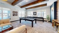 Located at 1003 148th St W in Rosemount, MN, this game room is a thoughtfully designed recreational space that blends rustic character with modern style. The ceiling is a standout architectural feature, adorned with bold, dark-stained reclaimed wood beams that contrast beautifully against the crisp white surround, lending warmth and visual depth to the room.

The main area is anchored by a sleek pool table and a full-length shuffleboard table, both finished in dark tones that complement the room's sophisticated palette. A large area rug grounds the space, while comfortable seating — including lounge chairs and a sofa — provides relaxed gathering areas for guests.

Natural light flows generously through multiple large windows, offering views of the surrounding neighborhood and brightening the neutral-toned walls. A bold teal accent wall adds a pop of color, enhanced by wall sconces and open wood shelving that provide decorative display space.

A striking fireplace clad floor-to-ceiling in a geometric patterned tile serves as a dramatic focal point in the lounge corner, topped by a mounted television. Nearby, a wood-paneled accent wall hosts arched decorative mirrors above a bar-height counter with gold stools, completing the room's inviting, upscale aesthetic.