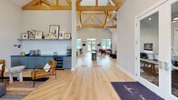 Located at 304 Battery Park Cir in Pendleton, SC, this beautifully designed single-floor space offers a generous, open layout that makes a striking first impression. The most dramatic architectural feature is the exposed natural wood timber truss system, which rises to soaring vaulted heights and lends the interior a warm, lodge-inspired character while filling the space with a sense of grandeur.

Light hardwood flooring runs throughout, complementing the neutral palette of soft white and gray walls. The open great room area is anchored by a bold floor-to-ceiling tiled fireplace wall, complete with a modern linear fireplace and wall-mounted display, creating a natural focal point for gathering. Adjacent seating areas are thoughtfully arranged, with curated artwork adding artistic energy to the space.

The kitchen area is outfitted with slate-gray shaker cabinetry accented with gold hardware, dark quartz countertops, a hexagonal mosaic tile backsplash, stainless steel appliances, and open floating shelves. Large windows along the kitchen wall allow abundant natural light to pour in.

A secondary lounge area features additional flexible seating beneath the exposed timber trusses, with large-scale contemporary artwork adorning the walls. The overall ambiance is modern yet inviting, blending industrial and artisan design elements seamlessly throughout.