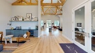 Located at 304 Battery Park Cir in Pendleton, SC, this beautifully designed single-floor space offers a generous, open layout that makes a striking first impression. The most dramatic architectural feature is the exposed natural wood timber truss system, which rises to soaring vaulted heights and lends the interior a warm, lodge-inspired character while filling the space with a sense of grandeur.

Light hardwood flooring runs throughout, complementing the neutral palette of soft white and gray walls. The open great room area is anchored by a bold floor-to-ceiling tiled fireplace wall, complete with a modern linear fireplace and wall-mounted display, creating a natural focal point for gathering. Adjacent seating areas are thoughtfully arranged, with curated artwork adding artistic energy to the space.

The kitchen area is outfitted with slate-gray shaker cabinetry accented with gold hardware, dark quartz countertops, a hexagonal mosaic tile backsplash, stainless steel appliances, and open floating shelves. Large windows along the kitchen wall allow abundant natural light to pour in.

A secondary lounge area features additional flexible seating beneath the exposed timber trusses, with large-scale contemporary artwork adorning the walls. The overall ambiance is modern yet inviting, blending industrial and artisan design elements seamlessly throughout.