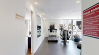 This fitness center is thoughtfully designed to support a comprehensive workout experience across interconnected zones. The main cardio area features dark rubber flooring and houses an array of treadmills, elliptical trainers, and stair climbers arranged along floor-to-ceiling windows that frame urban skyline views, flooding the space with natural light. Full-length mirrors along the walls enhance the sense of openness and allow users to monitor their form during exercise. Recessed ceiling lighting supplements the abundant natural light throughout.

A standout feature is the glass-enclosed strength training area, distinguished by a skylight ceiling that draws in daylight from above. This section is equipped with a multi-station cable machine, a flat bench, a spin bike, and a medicine ball, with large sliding glass doors opening to an adjacent outdoor terrace and pool area.

Adjacent to the fitness floor is a dedicated business center, finished with warm hardwood-style flooring and neutral walls. A long, minimalist workstation accommodates multiple users simultaneously, with ergonomic rolling chairs, ample power outlets, a wall-mounted monitor, and a built-in water fountain station nearby for convenience.