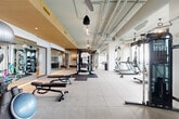 Located in the heart of Midtown Atlanta on Juniper Street, this contemporary fitness center spans a generously sized single floor, offering a comprehensive and well-appointed workout facility. The space is defined by its industrial-modern aesthetic, featuring exposed ductwork and piping across high ceilings, complemented by warm wood accents and stylish pendant lighting that balance the utilitarian elements with refined design.

The main workout floor is expansive and open, with large-format tile flooring and rubber mat zones accommodating a wide array of strength and cardio equipment, including treadmills, ellipticals, rowing machines, cable machines, power racks, free weights, and adjustable benches. Floor-to-ceiling windows line multiple walls, flooding the space with natural light and delivering sweeping panoramic views of the Atlanta skyline and surrounding greenery.

A dedicated studio or group fitness room features hardwood floors, mirrored walls, and a vibrant accent wall with a neon logo, opening directly onto an outdoor terrace with turf — ideal for yoga, stretching, or group classes with skyline backdrop views. A glass-enclosed private training room with a dark reflective ceiling offers an intimate setting for focused workouts. A hallway connects the various zones, with a small refreshment station featuring built-in cabinetry and a beverage cooler providing added convenience throughout the facility.
