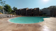 This outdoor pool area in Valley Park, Missouri, serves as a well-appointed communal amenity set against a backdrop of multi-story residential buildings. The centerpiece is a generously sized freeform swimming pool with clear, blue-green water, designed to accommodate a large number of swimmers comfortably. The pool's gently curved shape adds a leisure-oriented aesthetic to the space.

Surrounding the pool is an expansive stamped concrete deck finished in a warm, earthy tone, providing ample room for lounging and socializing. Rows of poolside lounge chairs and seating areas are arranged along the perimeter, offering plenty of space for sunbathing under open skies. Landscaped planters filled with colorful flowering plants and lush greenery are thoughtfully positioned throughout, adding natural vibrancy to the hardscape.

The area is fully enclosed by a metal security fence with a gated entry, and posted signage indicates that pool guests must be accompanied by a resident and that no diving is permitted. Mature trees surrounding the property provide partial shade and a sense of privacy. The multi-story residential buildings visible in the background feature balconies overlooking the pool, creating an integrated and cohesive outdoor living environment ideal for warm-weather recreation.