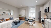 Located in the heart of Philadelphia on South Broad Street, this well-appointed single-floor residence offers a refined and cohesive interior design throughout its efficiently organized layout. The entry hallway sets an immediately elegant tone, featuring textured grasscloth-style wallcovering, light hardwood flooring, recessed lighting, and a curated gallery wall of large-scale framed artwork — creating a polished first impression.

The open living area seamlessly integrates a sitting space and dining nook. The living room is anchored by a plush sectional sofa, a black marble coffee table, and a large gold-framed mirror, complemented by a neutral blue area rug and warm wood media console. A generous window allows natural light to fill the room, enhancing the soft grey tones throughout.

The kitchen features floor-to-ceiling white shaker cabinetry, a white quartz countertop, a classic subway tile backsplash, and a full suite of stainless steel appliances including a range, dishwasher, refrigerator, and built-in microwave. A brushed gold faucet adds a refined accent detail.

The bathroom is a standout, showcasing a patterned black-and-white mosaic tile floor, a glass-enclosed walk-in shower with marble subway tile, matte black fixtures, and a warm wood vanity with a white countertop. The space is completed by a dedicated closet, providing practical storage.