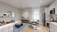Located in the heart of Philadelphia on South Broad Street, this well-appointed single-floor residence offers a refined and cohesive interior design throughout its efficiently organized layout. The entry hallway sets an immediately elegant tone, featuring textured grasscloth-style wallcovering, light hardwood flooring, recessed lighting, and a curated gallery wall of large-scale framed artwork — creating a polished first impression.

The open living area seamlessly integrates a sitting space and dining nook. The living room is anchored by a plush sectional sofa, a black marble coffee table, and a large gold-framed mirror, complemented by a neutral blue area rug and warm wood media console. A generous window allows natural light to fill the room, enhancing the soft grey tones throughout.

The kitchen features floor-to-ceiling white shaker cabinetry, a white quartz countertop, a classic subway tile backsplash, and a full suite of stainless steel appliances including a range, dishwasher, refrigerator, and built-in microwave. A brushed gold faucet adds a refined accent detail.

The bathroom is a standout, showcasing a patterned black-and-white mosaic tile floor, a glass-enclosed walk-in shower with marble subway tile, matte black fixtures, and a warm wood vanity with a white countertop. The space is completed by a dedicated closet, providing practical storage.