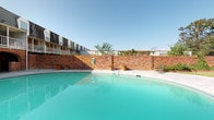 This single-floor residential unit offers a well-organized layout designed for comfortable everyday living. The open-concept living area is finished with plush wall-to-wall carpeting and neutral-toned walls, creating a bright and inviting atmosphere. A breakfast bar-style kitchen seamlessly connects to the living space, featuring dark cabinetry, black appliances, and a pass-through counter ideal for casual dining or entertaining. Hardwood-style flooring in the kitchen and hallway contrasts tastefully with the soft carpeting throughout the rest of the unit. The bedroom is a calm, carpeted retreat with a window dressed in horizontal blinds, allowing natural light to filter in while maintaining privacy. An electrical panel is conveniently accessible within the unit, and interior doors throughout are fitted with classic six-panel designs. The textured walls and ceilings add subtle architectural character to each room. Residents also enjoy access to a generously sized shared outdoor swimming pool, enclosed by a handsome full-height brick wall with lush climbing greenery. The pool area features a wide concrete surround and is framed by views of multi-story residential buildings under an open blue sky, offering a refreshing outdoor retreat.