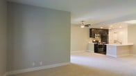 This single-floor space is designed with an open-concept layout that seamlessly connects the living area to the kitchen, creating a cohesive and functional environment. The living area features neutral-toned carpet flooring and soft gray walls accented by clean white baseboards, lending the space a calm, understated aesthetic. A ceiling fan is centrally positioned overhead, providing both lighting and air circulation throughout the shared space.

The kitchen area transitions naturally from the carpeted living space, featuring dark-toned cabinetry that contrasts with the lighter walls. Stainless steel appliances, including a range and an over-the-range microwave, are fitted within the cabinetry, offering a modern, practical finish. Recessed lighting in the kitchen area adds focused illumination, complementing the ceiling fan light in the adjacent living space.

The overall layout is straightforward and efficient, with minimal visual clutter and a neutral color palette throughout. The open flow between the living and kitchen areas enhances the sense of space, making the interior feel connected and easy to navigate. The design prioritizes functionality while maintaining a clean, contemporary appearance.