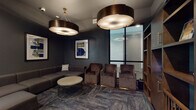 This single-floor amenity space combines upscale lounge design with an entertaining golf simulator experience. The lounge area is defined by deep charcoal and slate-toned walls that create a moody, sophisticated atmosphere, complemented by patterned carpet flooring in a herringbone-style layout. Seating is generous and thoughtfully arranged, featuring a large sectional sofa in warm grey upholstery alongside plush velvet accent chairs in rich brown tones, all centered around a round stone-topped coffee table with a metal wire base. Overhead, oversized drum pendant lights with bronze finishes serve as striking focal points, casting a warm, ambient glow throughout the lounge. Curated abstract artwork in bold blue and green hues adorns the dark walls, adding a contemporary artistic touch. Built-in wooden shelving provides both storage and display space, while glass-paneled doors connect the lounge to an adjoining corridor, maintaining an open and accessible flow. The golf simulator room is a dedicated, darkened space with matte black walls and a full-scale projection screen, complete with a synthetic turf hitting mat. Decorative circular canvas art adds personality to this entertainment zone. Natural light filters into the lounge through large windows, offering a balanced contrast to the intentionally dramatic interior palette.